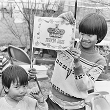 A vintage black and white photo shows three Vietnamese children smiling to the camera while holding up Mardi Gras souvenirs including a flag.