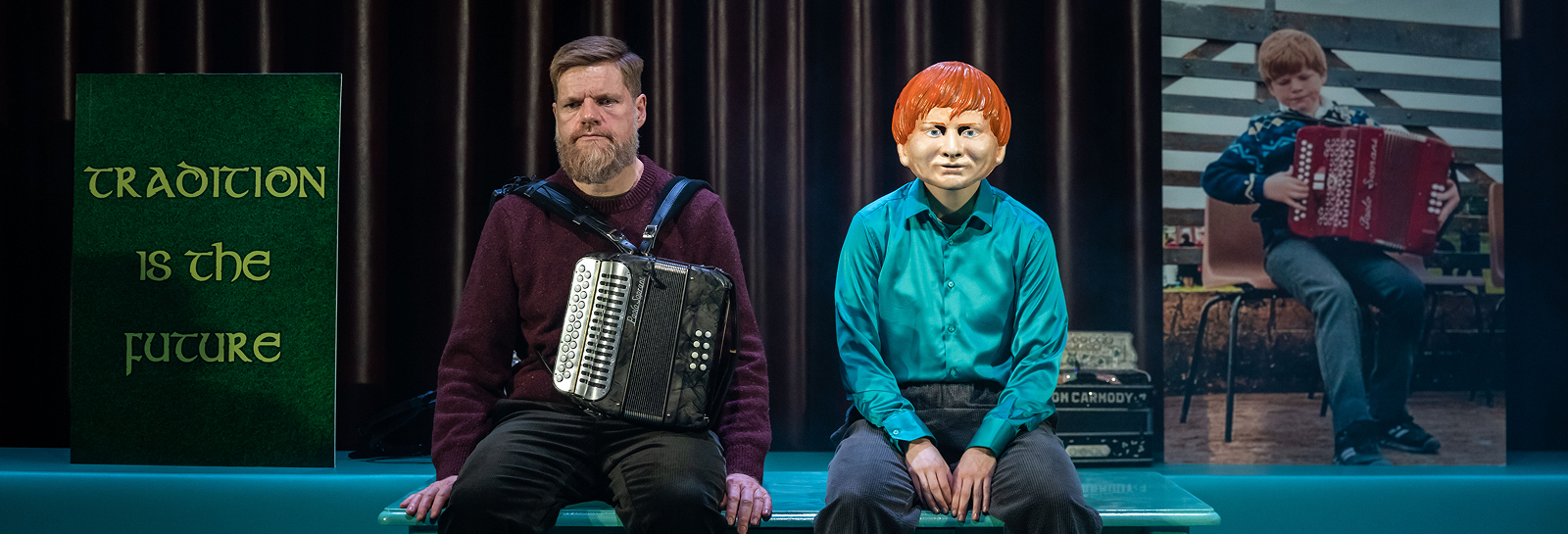 An image of two people on stage. One of them stands looking into a small telescope with his other arm outstretched. The other sits in an armchair with round sunglasses on. 