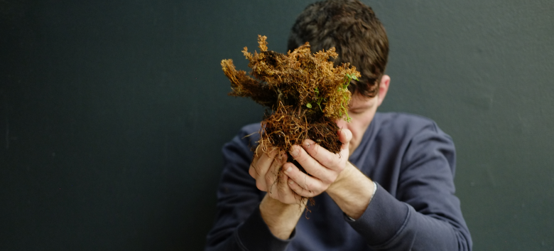 A person in a navy sweater holding a piece of moss. He cups it with both hands, and holds it in front of his face. 