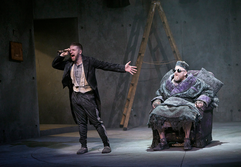 An image of two people on stage. One of them stands looking into a small telescope with his other arm outstretched. The other sits in an armchair with round sunglasses on. 