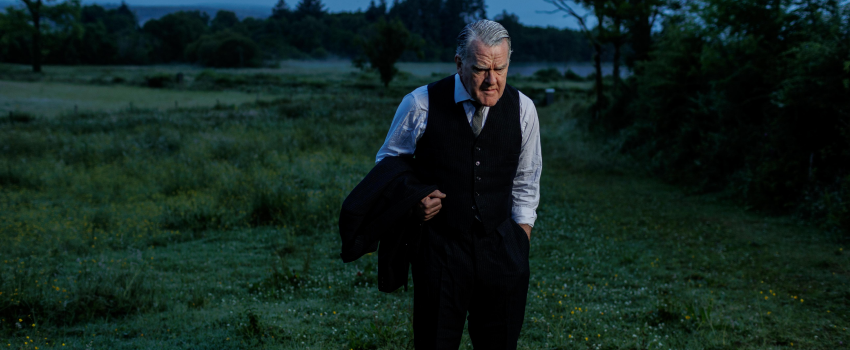 A man stands outside in a dark field. He wears a pants, waistcoat, shirt and tie, and carries his jacket over his arm. He has a solemn look on his face. 