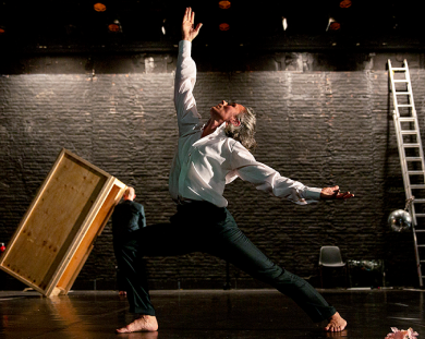 A person on stage wearing a white shirt and black pants. They are barefoot and holding an outstretched pose.