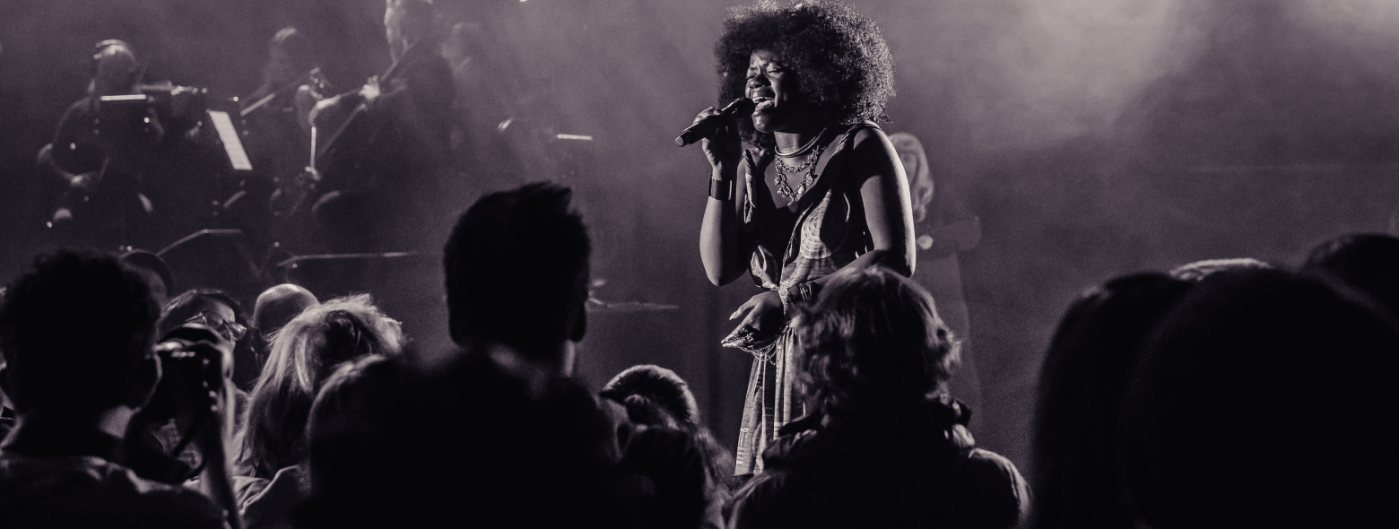 A black and white image of the artist Winnie Alma singing in front of a standing audience. An accompanying band plays onstage behind her.