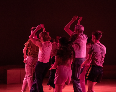 A group of dancers in casual clothing facing away from the camera. Some have their arms loosely by their sides and some hold them over their heads. 