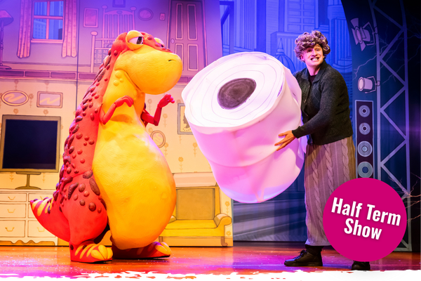 A person in a dinosaur costume, and a person dressed as an old lady stand on a stage holding a comically large toilet roll.