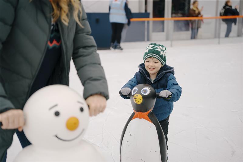 A boy is a warm hat and gloves holds onto a penguin to help him stake