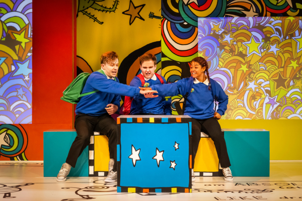 Three people dressed as school children stand on a stage with their hands together in celebration