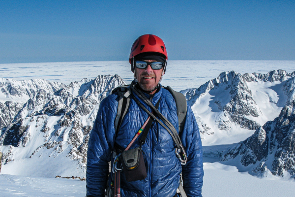 A man in a blue jacket and red helmet stands with snow-covered mountains in the background