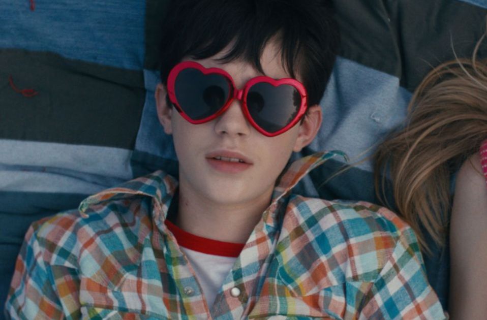 A close up of a young person, lying down looking at the sky with oversized red heart shaped sunglasses on. 