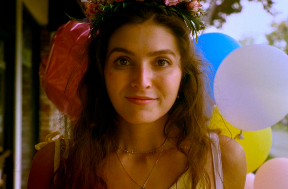 Close up of a young woman, smiling, with a flower wreath in her hair and colourful balloons behind her. 