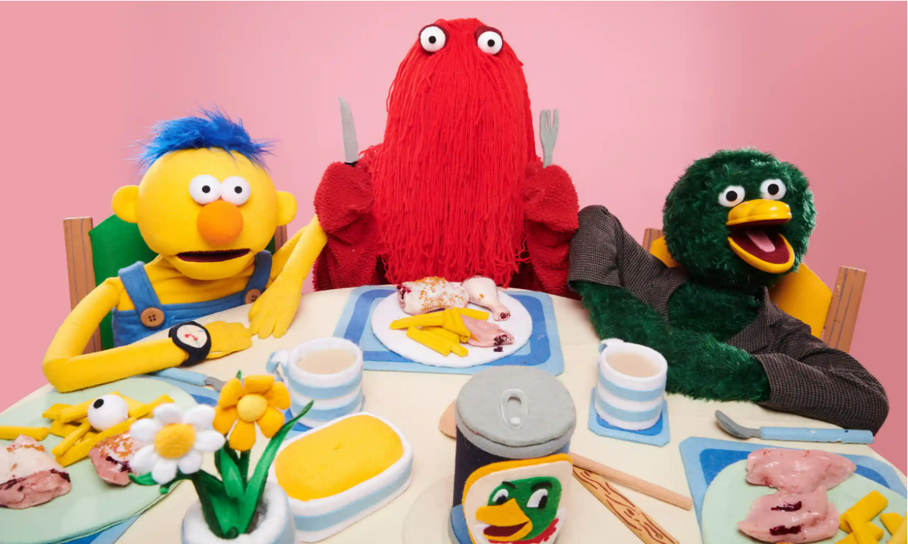 Several puppets sit around a dinnertable, a yellow puppet with blue hair, a red puppet that looks like a mop-head and a green duck. 