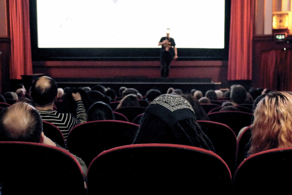 An audience in a cinema looks at the figure at the front of the stage. 