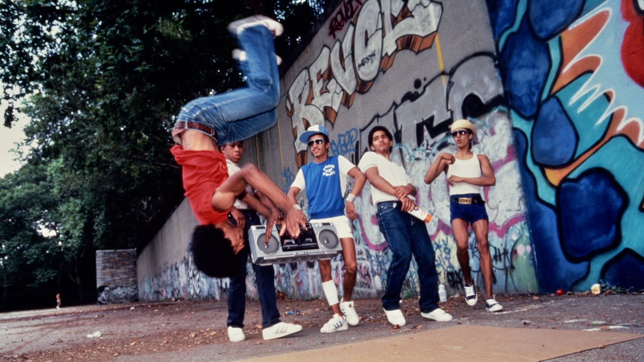 A still from the film 'Wild Style' showing young people in hip-hop style clothing dancing on the street. A black man with an afro wearing a red t shirt, blue jeans and white trainers is captured upside-down performing a backflip, in front of a wall covered in graffiti art.