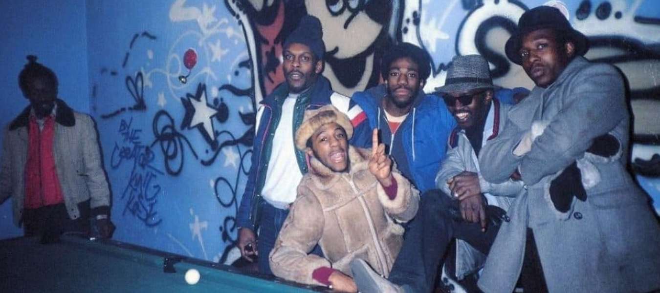 A still from the film 'Wild Style' showing a photograph of several black, male musicians wearing fur coats and hats, gathered around a pool table.