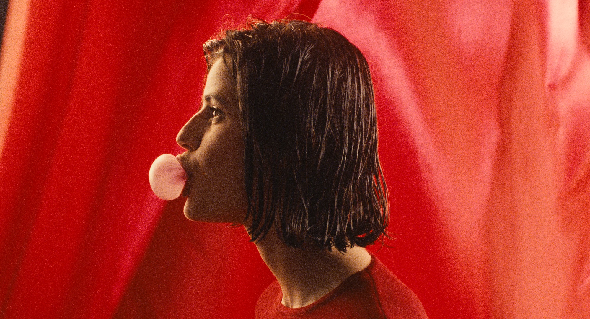 A profile show of a  young woman blowing bubblegum, in front of a red fabric backdrop. 