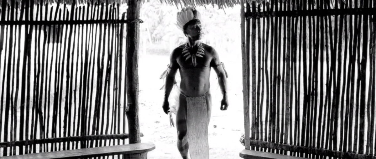 A black and white still of a man dressed in tribal clothing, walking through a doorway into a room made of loose sticks.