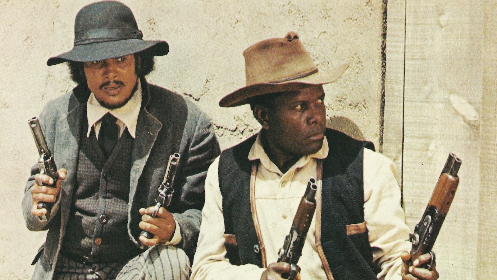 A still from the film Buck and the Preach: Two men dressed in cowboy era clothing lean against a wall, old pistols in each hand. 