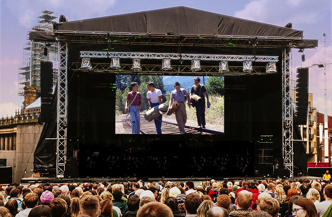 A crowd of people sit watching a big screen erected on a concert style stage, outdoors.