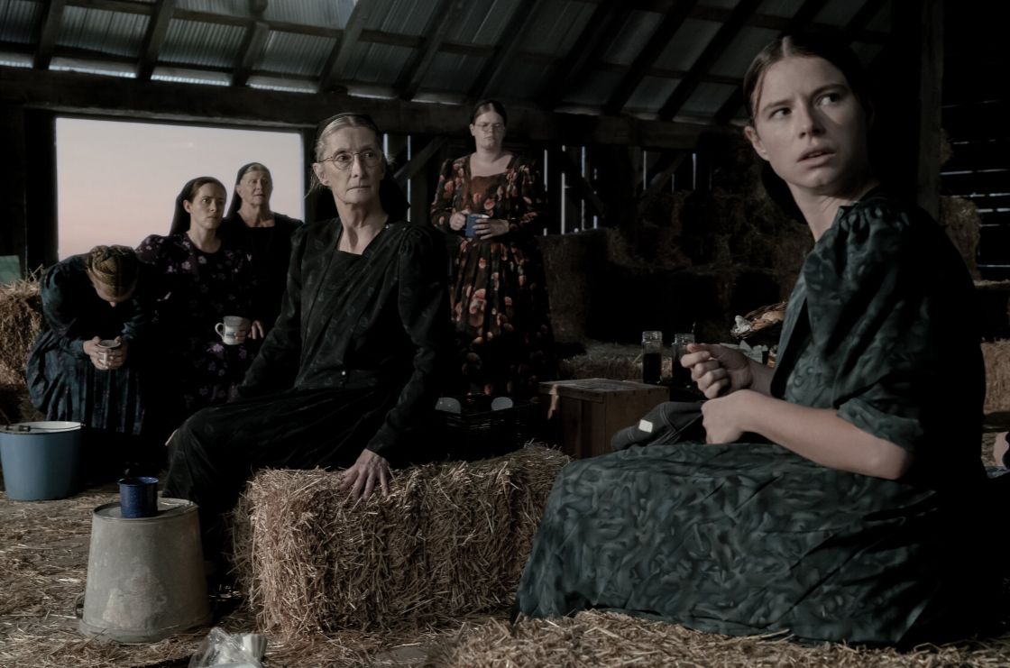 A group of women dressed in old fashioned smocks gather together in a barn, sat on bales of hay.