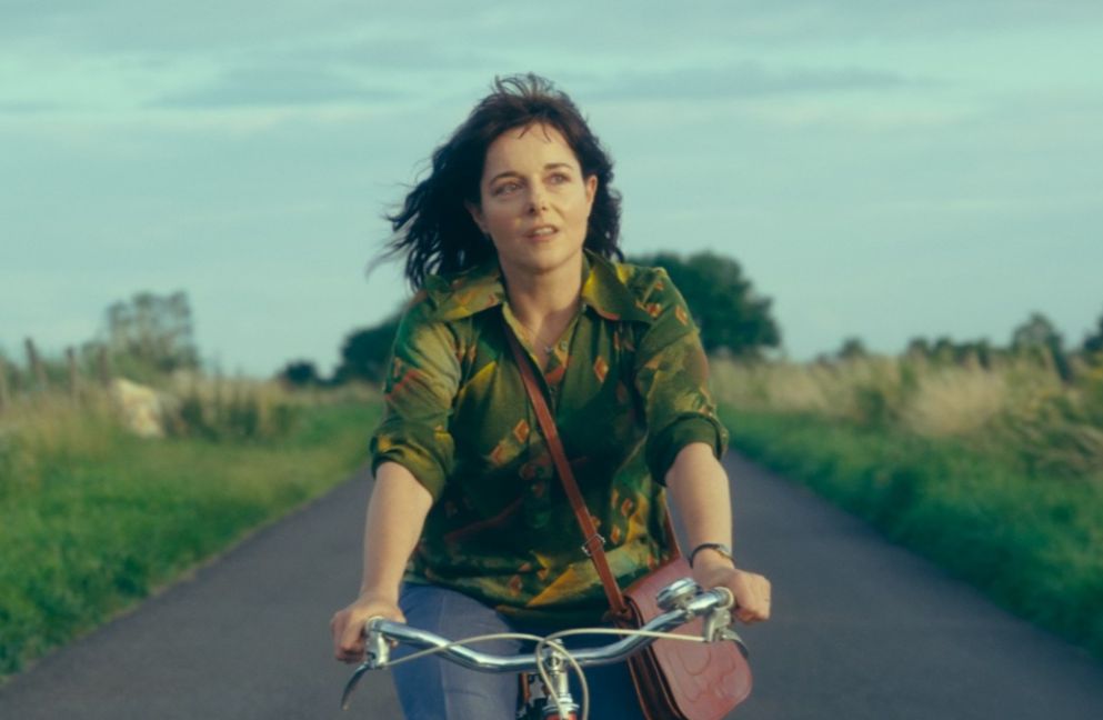 A woman cycles in the middle of a small road, surrounded by fields, looking at something in the distance. 