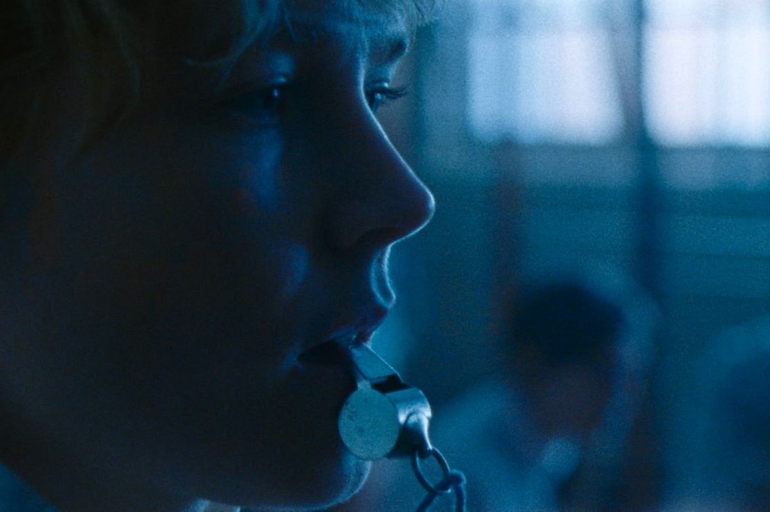 A close up of a woman's face in profile, a whistle in her mouth, everything bathed in a dark blue light. 