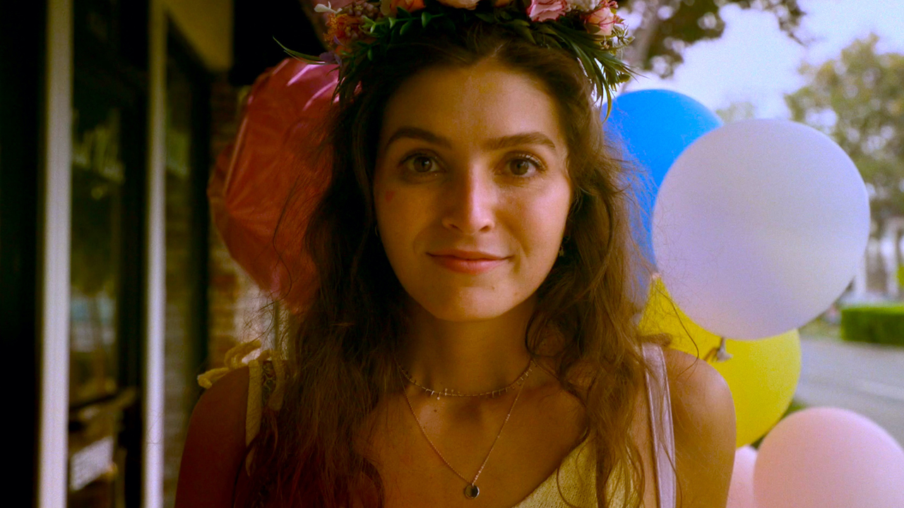 Image of a young woman, smiling directly at you, with balloons and a colourful wreath around her head. 