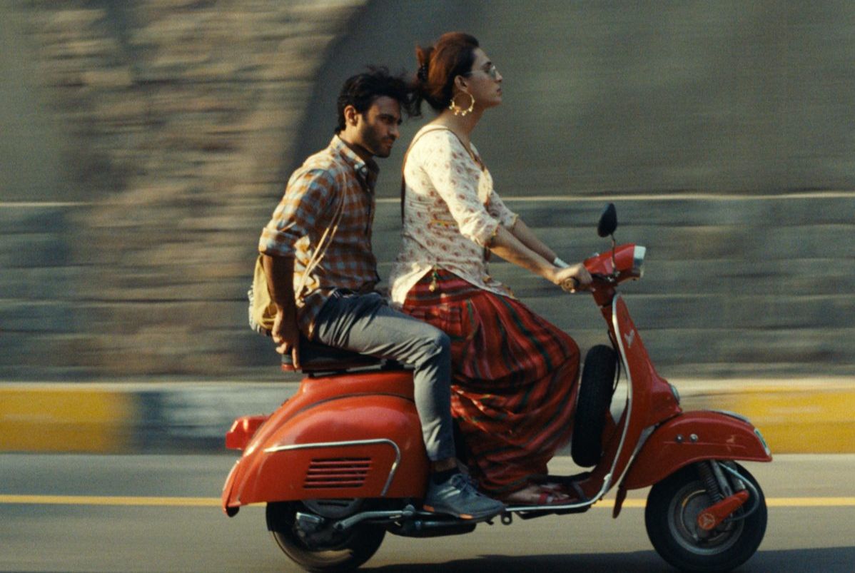 A man sits on the back of a retro moped, driven by a young woman in sunglasses. 