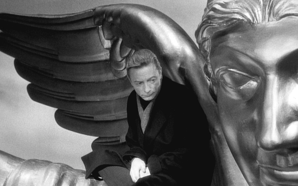 A black and white still from a film where a man in a suit sits on top of a large metal angel.