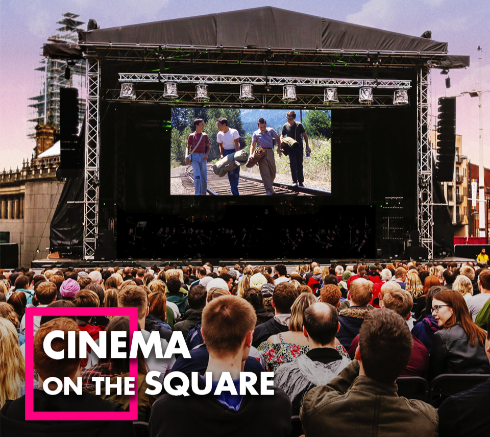 A crowd stands in front of a big stage with a large screen on it, in front of a dusky sky. A movie plays showing four boys walking down a trainline. TEXT: Cinema in the Square