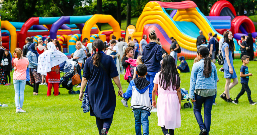 A family hold hands through a park full of other families, inflatables can be seen in the distance. 