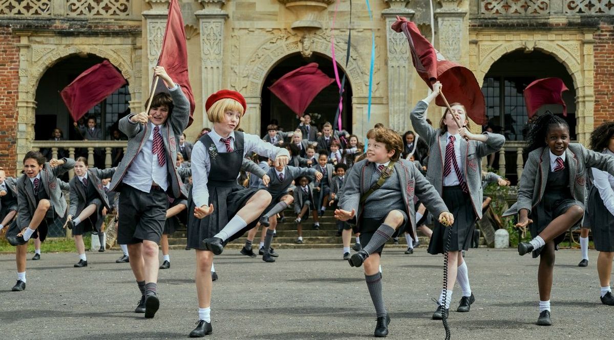 Several schoolchildren dance and sing with flags in front of their school. 
