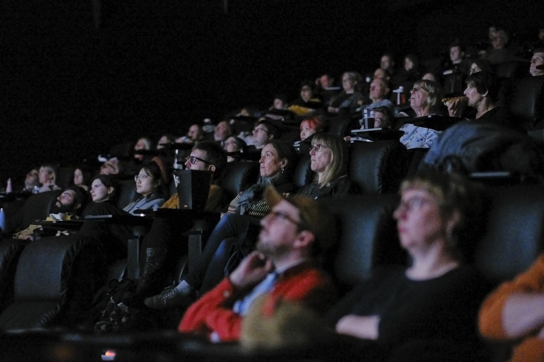 An audience sits in a cinema, their faces all lit up by the glow of the screen. 