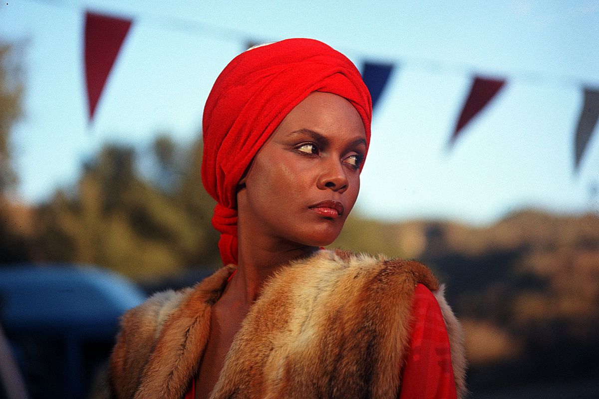 A black woman in a fur coat and red headwrap stares off into the distance. 
