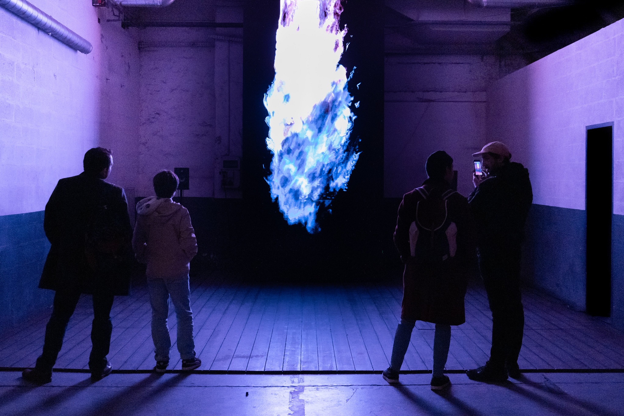 A group of people stand in front of a  intense, large blue flame on a floor length black screen