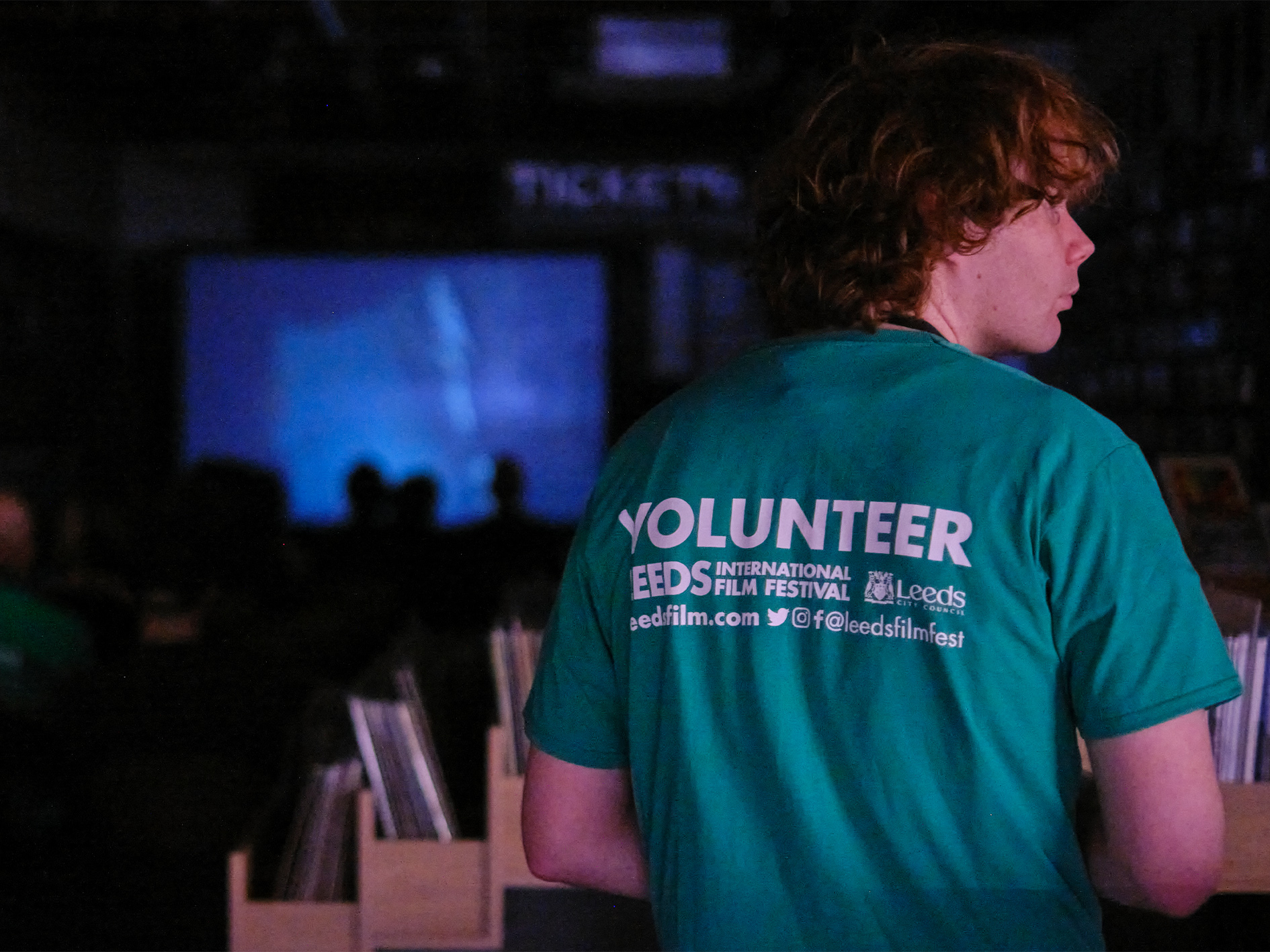 A young person can be seen from behind with "VOLUNTEER" written on their T-Shirt. A screen with the silhouette of an audience can be seen in the background. 
