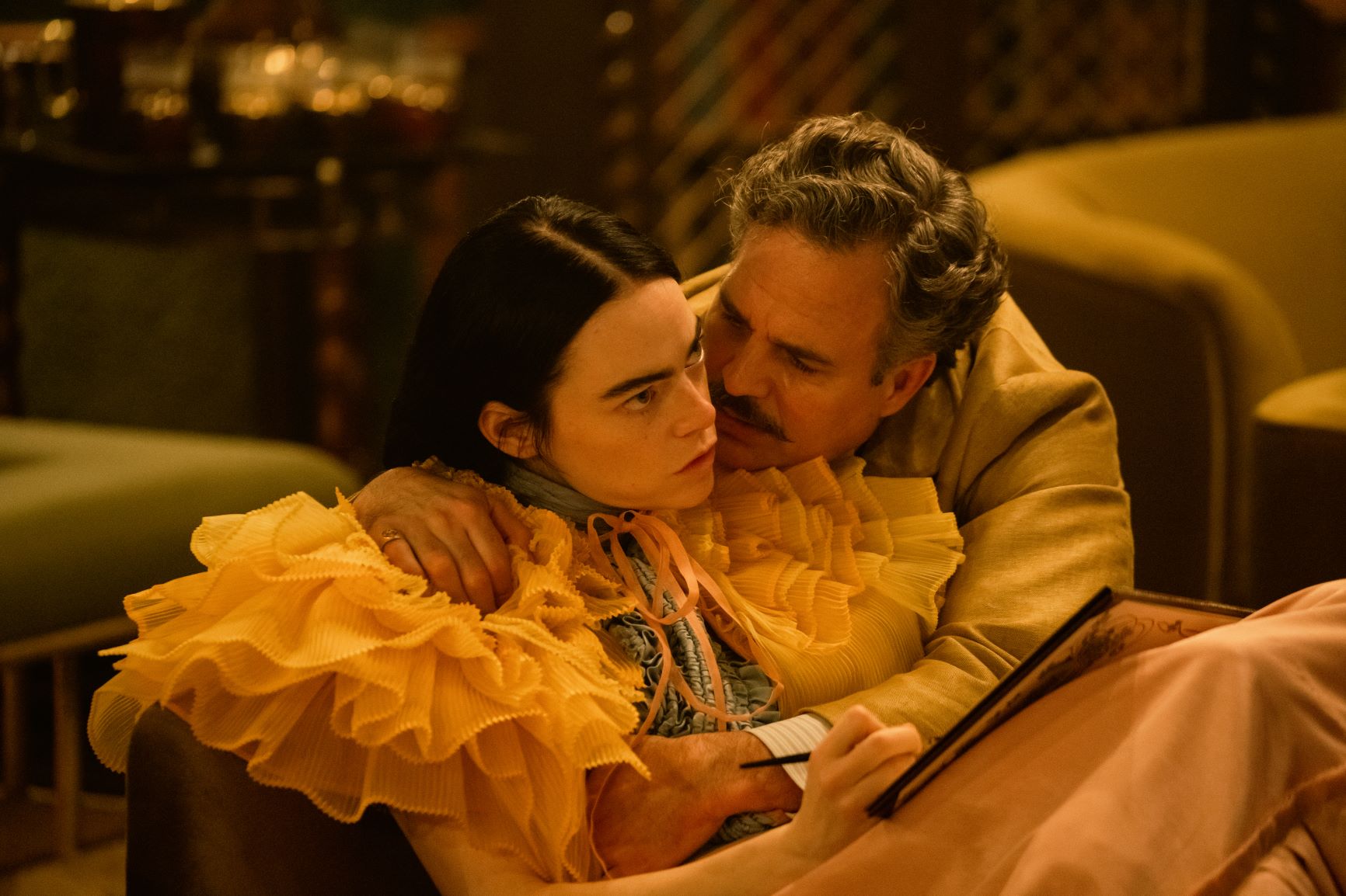 A woman with thick eyebrows and a frilly outfit, stares off intensely while a mustached man cuddles against her. 