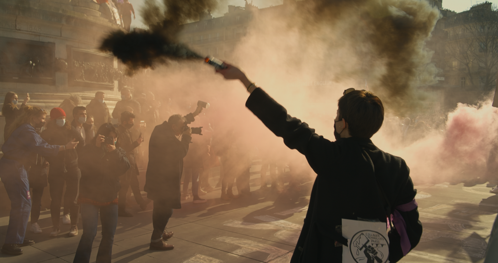 Smoke covers a group of people, many are wearing masks and have camera. The back of a protester can be seen in the foreground, waving a smoke device. 