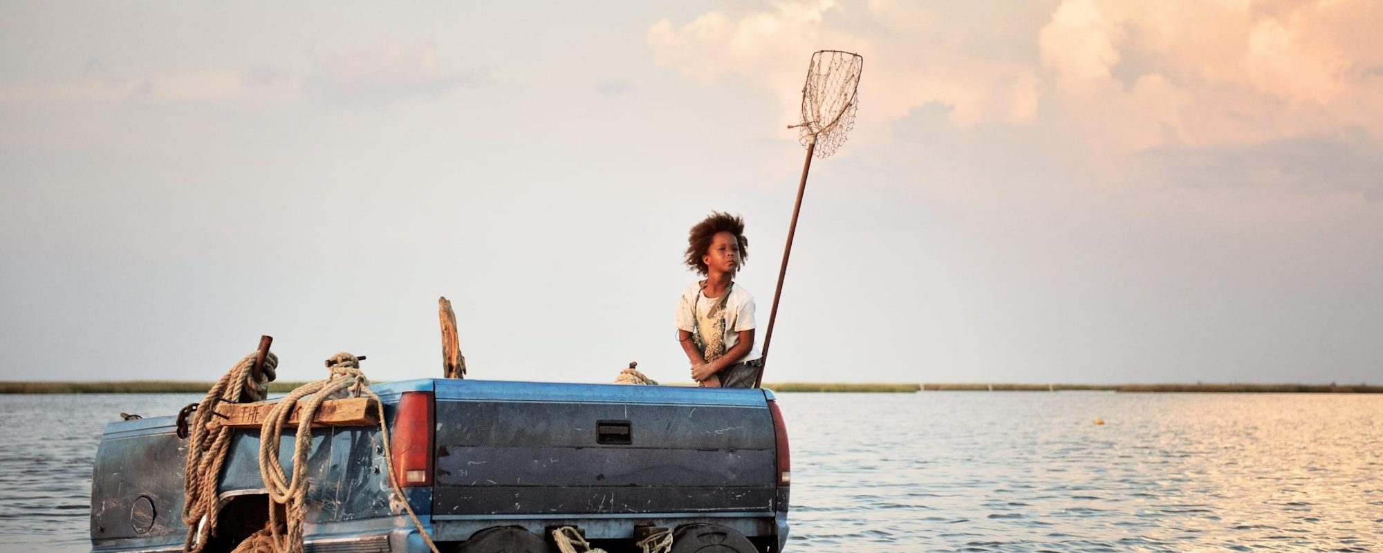 A young girl rides the back of a truck like a boat, through a mass of water, with a fishing net next to her, poking up into the sky. 