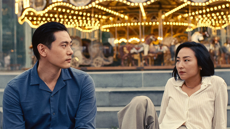 A man in a casual dark blue shirt, looks into the eyes of a casually dressed woman sat next to him. Behind them, you can see the gold lights of a carousel. 