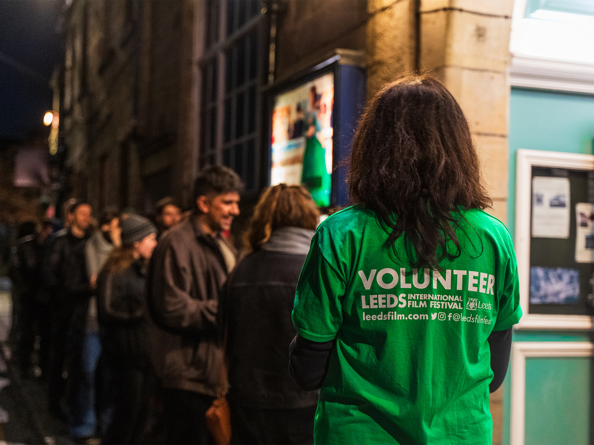 A young person can be seen from behind with "VOLUNTEER" written on their T-Shirt. A line of people queuing up for a venue stand in front of them. 