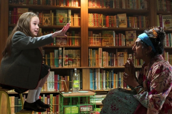 A young girl in school uniform with her hand outstretched facing a woman in patterned clothes drinking from a bottle. They are mid-conversation.
