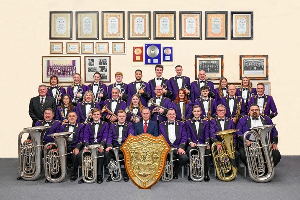 Members of the Brighouse & Rastrick Band in purple concert dress stood and sat in rows with their instruments.