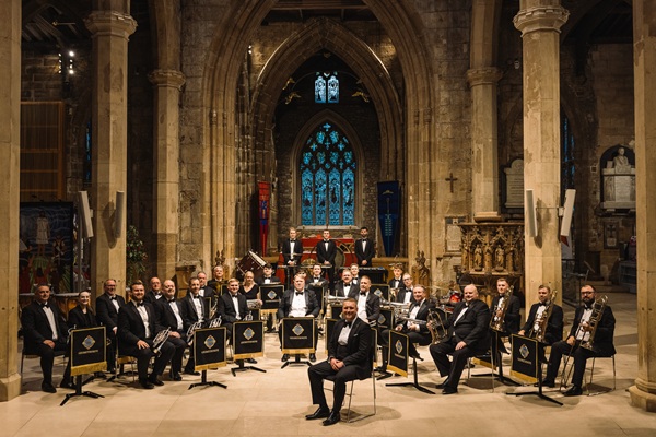 Members of the Grimethorpe Colliery Band in black concert dress sat in formation with their brass instruments in a church.