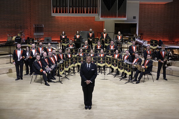 Players of Black Dyke Band in black and red concert dress in formation on stage with their instruments.