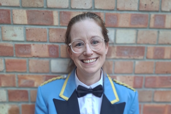 A young woman wearing glasses and blue suit concert dress against a brick wall.