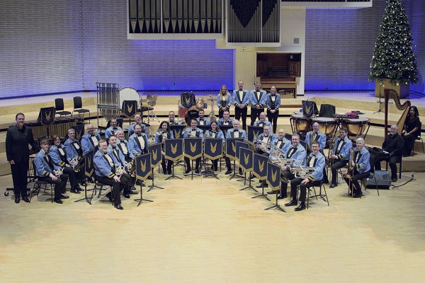 Members of the KNDS Fairey Band in concert dress in a concert hall with their instruments.