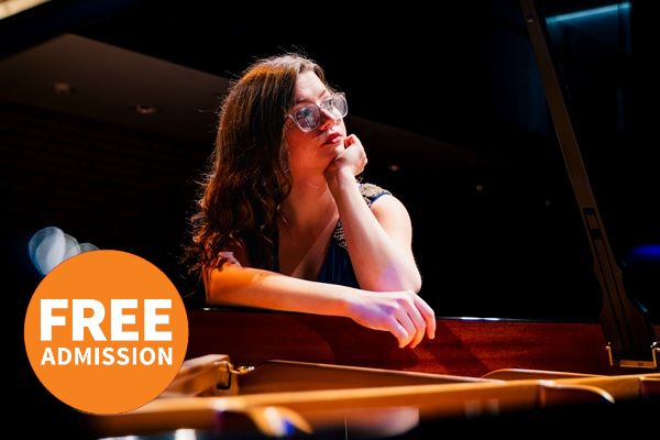 Pianist Madeleine Brown wearing clear acetate framed glasses and a sleeveless top. Madeleine is leaning on the piano with one hand on her face and the other over the piano.