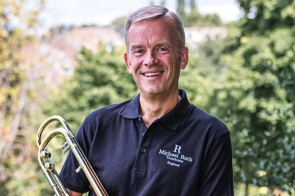 A man in a dark coloured polo shirt smiling with his trombone.