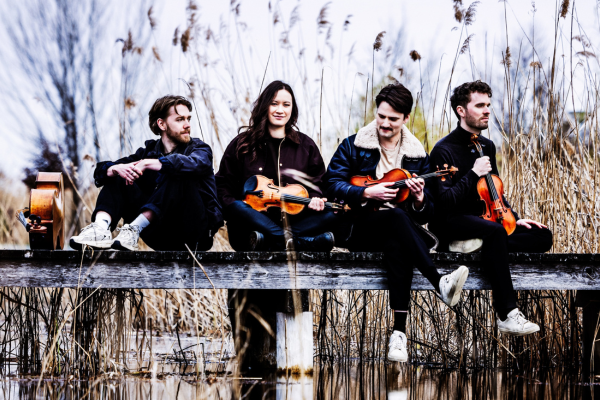 The four members of Opus13 in casual clothes sat on a wooden walkway pier in a nature reserve with their string instruments.