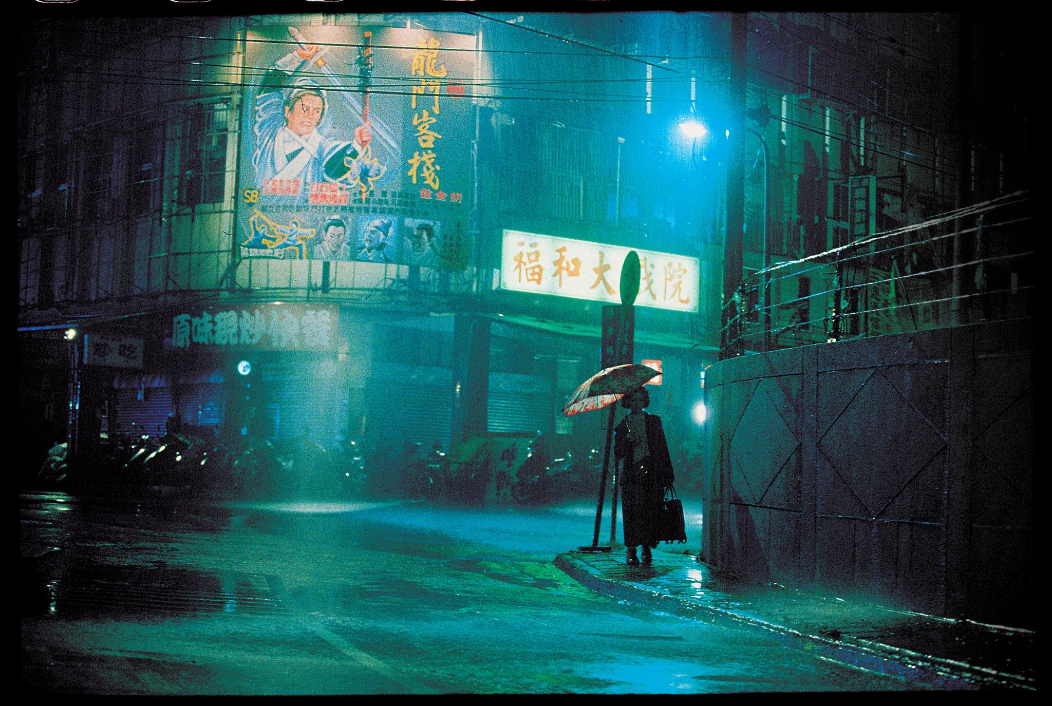 A  figure in the distance stands with an umbrella, blue street light illuminating the streets. 